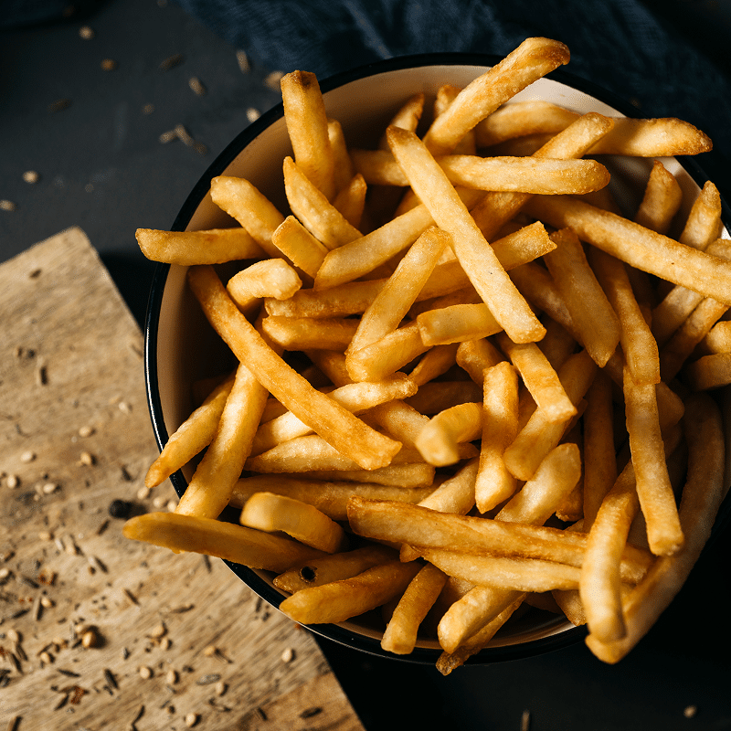 Gros plan sur des frites dorées croustillantes servies dans un bol blanc à bord noir, sur fond sombre et planche en bois épicée.